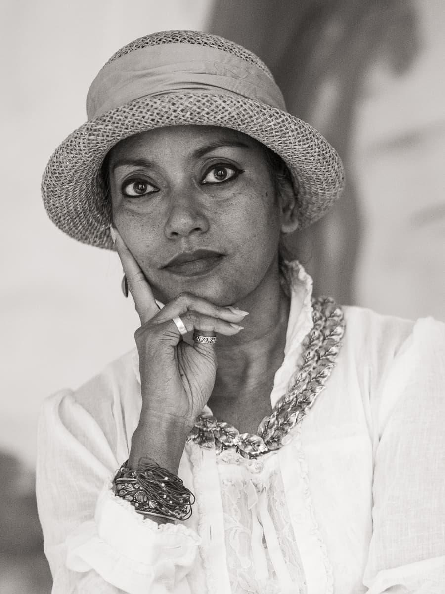 Black-and-white portrait of Murshida Arzu Alpana in a woven straw hat and white lace blouse, her chin resting on her hand